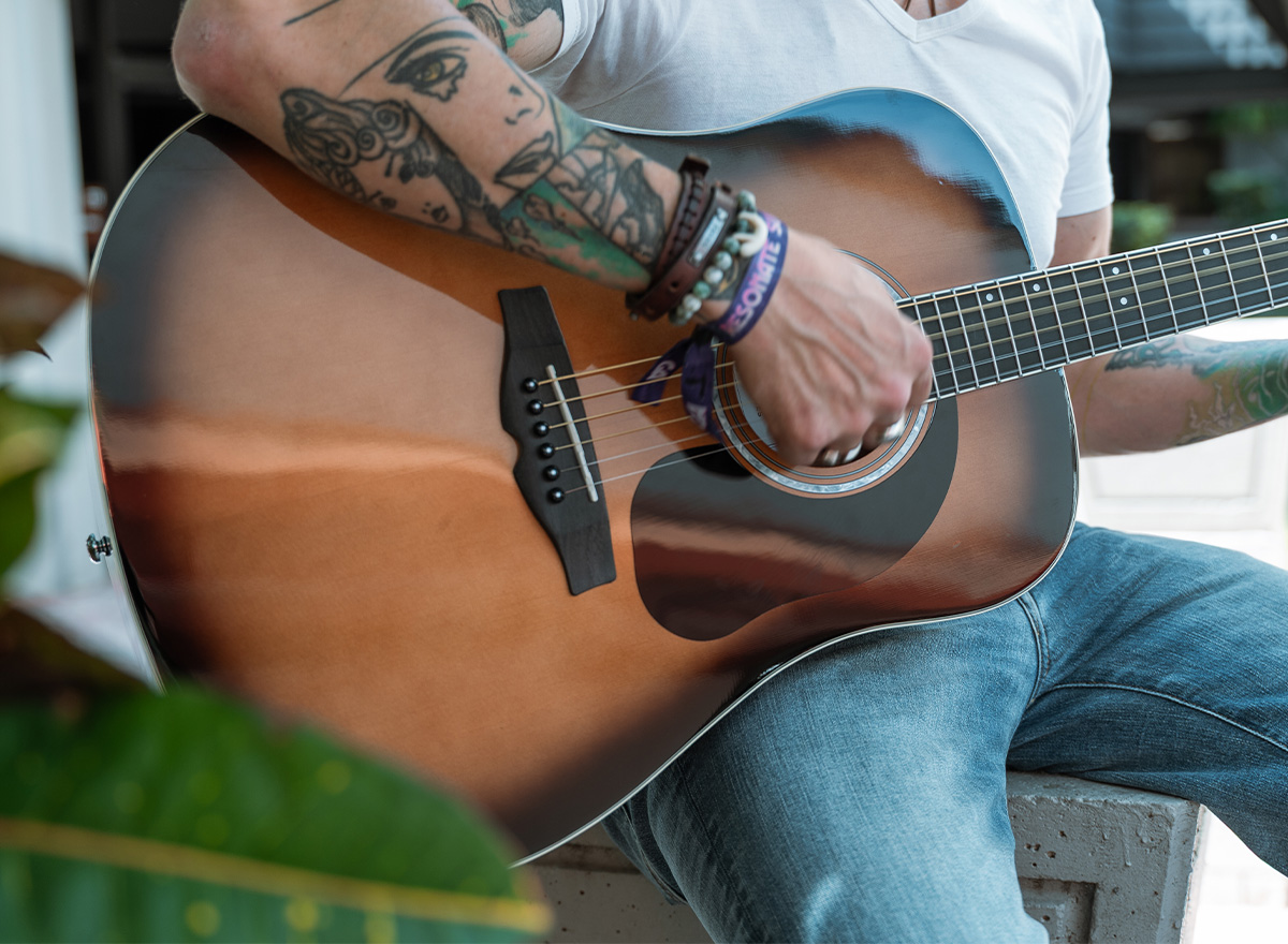 Tattooed musician strumming sunburst Mitchell acoustic guitar outdoors